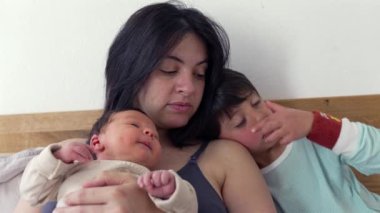 Anne bebeği kollarında tutarken, bileklikli büyük kardeş onun yanında oturup gözlerini ovuşturuyor. Sahne, aile hayatının hassas bir anını yakalar. Paylaştıkları bağı ve yakınlığı gösterir.