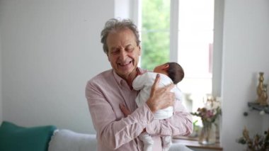 Büyükbaba yeni doğmuş bebeğe sevgiyle bakıyor, parlak bir oturma odasında bebeği kucağında tutuyor, büyük anne ve büyük baba ve torun arasındaki sevgi ve bağları gösteriyor.