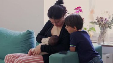 Anne, büyük oğlu izlerken yeni doğan bebeği koltukta emziriyor. Evde aile bağları, doğal ışık ve rahat, sevgi dolu bir atmosfer.
