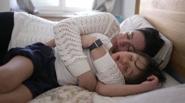 Anne ve küçük çocuk yatakta uzanıyor, kucaklaşıp huzur içinde uyuyor, aile bağlarının rahatlığını ve yakınlığını vurguluyor, ebeveyn-çocuk ilişkilerine odaklanmış yaşam tarzı için ideal.