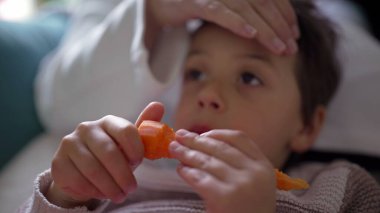 Atıştırmalık yiyen genç bir çocuğun yakın çekimi, düşünceli bir yüz ifadesi, anne arka planda, samimi aile anı, oturma odası düzeni, yemekten zevk alan çocuk, ev sahnesi, odaklanmış görünüm.