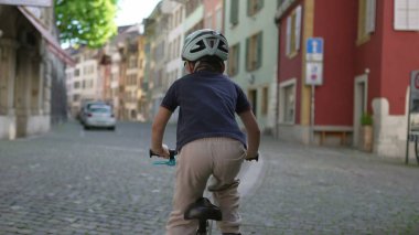 Kasklı bisikletli bir çocuk renkli binaları olan eski şehir caddesinde geziyor, tarihi şehir ortamlarında açık hava maceraları sergiliyor ve aktif bir çocukluk geçiriyor, yaşam tarzı görüntüsü için ideal.