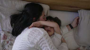 Anne ve küçük çocuk yatakta yatıyor, anne çocuğun alnından nazikçe öpüyor, huzurlu ve sevgi dolu bir aile anını yakalıyor, ebeveyn-çocuk ilişkisine odaklanmak için ideal bir yaşam tarzı.