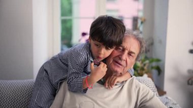 Küçük bir çocuk, büyükbabasına sarılıp bir şeyler atıştırıyor. Evdeki bir kanepede sevgi ve yakınlık paylaşıyor.