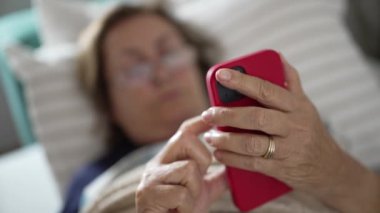 Yatakta yatarken kırmızı akıllı telefonuyla meşgul olan yaşlı kadın, huzur ve rahat bir ortamda teknolojiye uyum sağlayan yaşlı vatandaşı temsil ediyor.