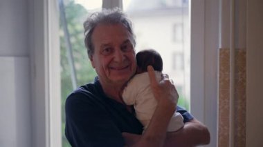 Pencerenin yanında bebek tutan yaşlı bir adam, sevgi ve neşe ifade ediyor, nesiller arası bağ ve aile anlarını bir ev ortamında vurguluyor.