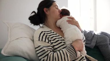 Anne, yeni doğmuş bebeği nazikçe kucağında tutarken genç çocuk seyrediyor sıcak ve sevgi dolu bir yuva ortamındaki aile bağını vurguluyor.