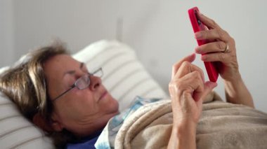 Yaşlı bir kadın kırmızı akıllı telefonunu kullanarak rahat bir şekilde yatakta uzanıyor, rahat bir ev ortamında teknolojinin ve yaşlıların kesişiminin altını çiziyor.