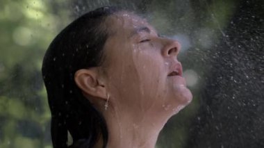 Gözleri kapalı bir kadın, yavaş çekimde ferahlatıcı bir duşun tadını çıkarıyor. Yüzüne su damlacıkları düşerek huzurlu ve huzurlu bir anı yakalıyor.