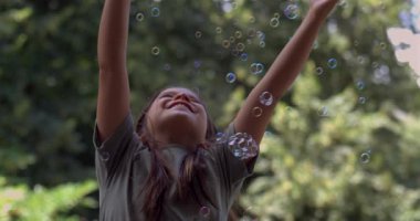 Kolları havada ve başı arkaya eğik bir kız, 800 fps 'te ağır çekimde kabarcıkların tadını çıkartıyor, güneşli bir günde neşeli ve neşeli bir an yaşıyor.
