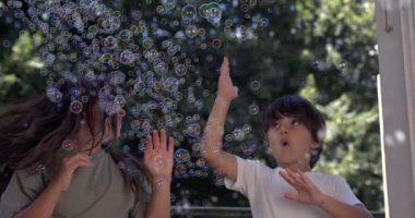 Genç kız ve oğlan 800 fps 'de baloncukların keyfini çıkarıyorlar, etraflarında kabarcıklar uçuşuyor, keyifle uzanıyor ve açık havada gülüyorlar.