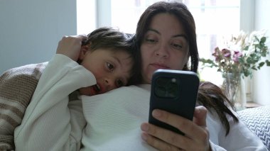 Anne ve çocuk kanepede kaynaşıyor, akıllı telefon kullanıyor, aile zamanlarını ve modern teknolojiyi ev ortamında sergiliyor, bağlantıyı ve rahatlığı vurguluyor.