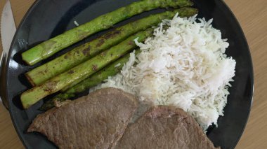 Izgara kuşkonmaz, taze biftek ve yumuşak beyaz pirinç tabağı. Beslenme ve yemek fotoğrafçılığı için mükemmel dengeli bir yemek sunuyor. Sağlıklı bir akşam yemeği seçeneğini sergilemek için ideal.