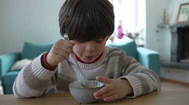 Genç çocuk masada bir kase yoğurt yiyor, kaşığı düşünceli bir ifadeyle ağzına doğru kaldırıyor, ev ortamında sakin ve samimi bir anı yakalıyor.