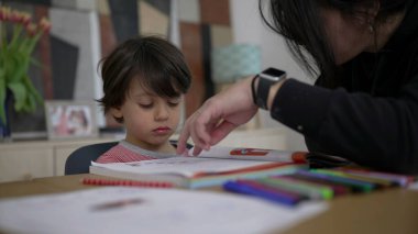 Anne, küçük çocuk ev ödevi sırasında hayal kırıklığına uğramış ve isteksiz hissederken, destek öneriyor. Masada renkli eğitim materyalleri, evde öğrenme konusunda zorluklar ve teşvik sergiliyor.