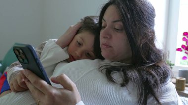Anne, çocuğu koltukta rahatlatıyor, akıllı telefon kullanıyor, modern evlerde aile bağlarının ve teknolojinin önemini vurguluyor, rahatlatıcı bir ortamda bakım ve yakınlığı vurguluyor.