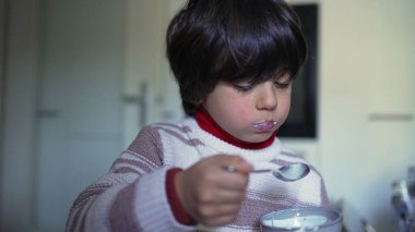 Genç çocuk kaşıkla yoğurt yiyor, mutfakta neşeli ve neşeli bir yüz ifadesi sergiliyor, keyifli ve spontane bir çocukluk anını tasvir ediyor.