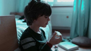 Çocuk bir parça ekmek yiyor, sessiz bir atıştırma anının tadını çıkarıyor, aydınlık bir odada oturuyor, sakinlik ve memnuniyeti yansıtıyor.