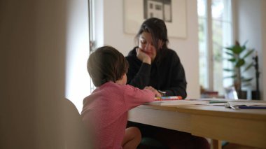 Çocuğun arkasından bakıldığında, çocuk ve ebeveyn bir masada öğrenme anını paylaşıyor ve bir ev ortamında ebeveynlerin eğitime katılımının önemini gösteriyor.