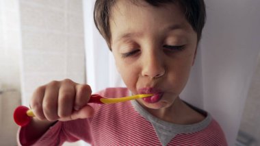 Dişlerini renkli bir diş fırçasıyla fırçalayan, çizgili pijama giyen, iyi aydınlatılmış bir banyoda ağız temizliği alışkanlıkları ve çocukluk rutinlerine odaklanan genç bir çocuk.