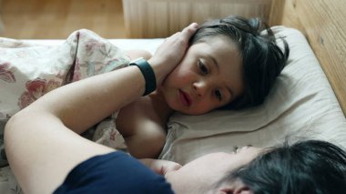 Anne, oğulların yüzünü yatakta okşar, rahat bir sabah rutini, ev yatak odası, samimi aile anı, sıcaklık ve sevgi, bağ kurma zamanı, günün rahat başlangıcı, çocuk gülümsemesi. 