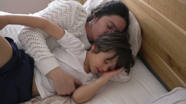 Anne ve çocuk yatakta huzur içinde uyuyor, huzurlu ve samimi bir dinlenme ve kaynaşma anı yakalıyorlar, paylaştıkları sevgi ve rahatlığı vurguluyorlar.