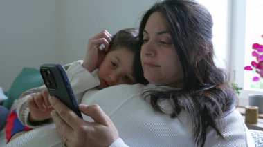 Anne, çocuğu koltukta rahatlatıyor, akıllı telefon kullanıyor, modern evlerde aile bağlarının ve teknolojinin önemini vurguluyor, rahatlatıcı bir ortamda bakım ve yakınlığı vurguluyor.