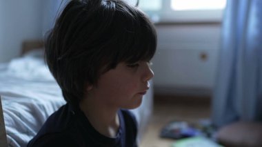 Koyu renk saçlı bir çocuğun profili, sakince oturuyor, içsel ve melankolik görünüyor, yumuşak ışıklı bir odada sessiz bir anın tadını çıkarıyor.