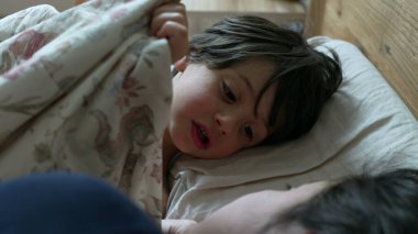 Çiçekli battaniyenin altında yatan genç çocuk, sabah rutini, ev yatak odası, günün konforlu başlangıcı, aile anı, uyanma, neşeli yüz ifadesi, rahat ve sıcak, evcil sahne