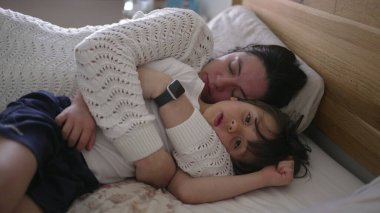 Anne ve küçük çocuk yatakta yatıyor, anne başını çocuğun göğsüne yaslıyor, huzurlu ve samimi bir aile anını yakalıyor, aile sevgisini ön plana çıkaran yaşam tarzı için mükemmel.