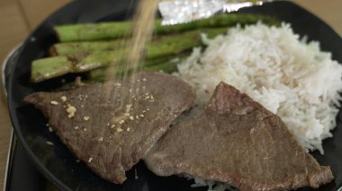 Siyah tabakta ızgara kuşkonmaz ve beyaz pirinçle servis edilen tipik bir Brezilya malzemesi olan farofa ile süslenmiş biftek, yemek fotoğrafçılığı için ideal ve lezzetli bir yemeği vurgular.