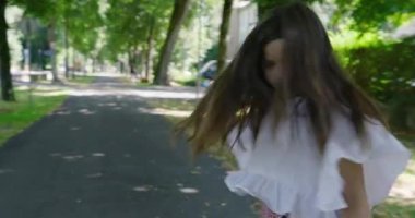 Uzun saçlı bir kız neşe içinde ağaçlarla çevrili güneşli bir parkta ağaçlarla ve yeşilliklerle çevrili ağaçlarla çevrili ağaçlarla çevrili ağaçlarla çevrili bir yolda dönüyor, özgürlük ve mutluluk hissi yaratıyor.