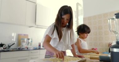 Mutfakta kardeşler yemek hazırlıyor, kız ve oğlan yemek pişirmeye odaklanıyor, aşçılık faaliyetleri, aile ilişkileri, arka planda anne, ışık dolu mutfak.