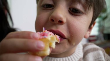 Çocuk büyük bir ısırık alır pembe şekerli donuttan, renkli serpilmiş, çocukluk hoşgörüsünü ve heyecanını temsil eden, ağzının ve ellerinin kapalı çekimini,