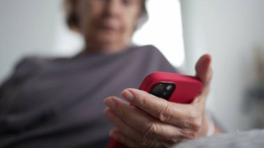 Evdeki bir kanepede oturmuş internetteki içeriği okumakla meşgul son sınıf bir kadının cep telefonunu tutarken yakın plan fotoğrafı.
