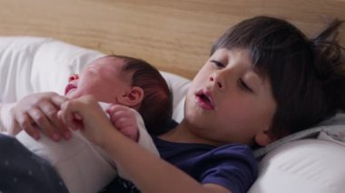 Küçük çocuk yeni doğan kardeşini yatakta nazikçe tutuyor, sakin bir ifadeyle kameraya bakıyor, tatlı ve koruyucu bir kardeş anı yakalıyor.