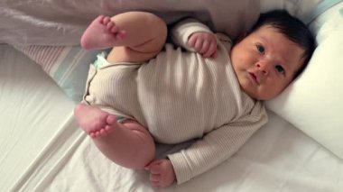 Yeni doğan bebek yatağa uzanıyor, meraklı gözlerle bakıyor, çizgili bir tulum giyiyor, rahat, huzurlu bir ortamda yumuşak bir yatak örtüsüyle çevrili.