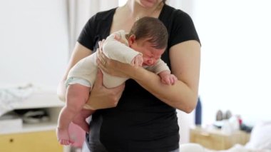 Ağlayan yeni doğmuş bir bebek, çizgili bir tulum giymiş bir anne tarafından tutuluyor, hafifçe aydınlatılmış bir odada bir anlık bakım ve rahatlık taşıyor.