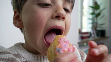 Genç bir çocuk pembe şekerli donut yiyor. Renkli serpilmiş, çocukluk hoşgörüsünü ve neşesini temsil ediyor. Yüzüne ve ellerine yakın plan, kapalı alan.