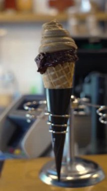 Çikolatalı ve vanilyalı leziz dondurma külahı, rahat bir kafe içinde modern bir tutucunun içinde sergileniyor, hoşgörü ve tatlı bir hazzı çağrıştırıyor.
