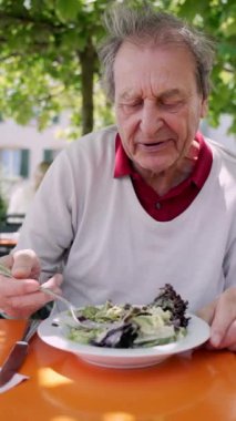 Açık bir kafede taze salata yiyen yaşlı bir adam, dingin ve yeşil bir ortamda sağlıklı bir yemeğin tadını çıkarıyor, beslenme ve refahın önemini vurguluyor.