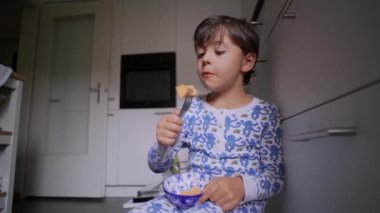 Çocuk mutfak zemininde oturmuş çatalla sağlıklı bir kavun atıştırıyor, tatmin olmuş bir şekilde gülümsüyor.