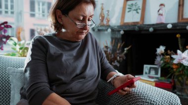 70 'li yaşlarda, oturma odasındaki koltukta rahat bir şekilde otururken cep telefonu kullanan, okuma gözlüğü takan, telefonu tutan, burnunu tırmalayan ve gözlüğünü çıkartan yaşlı bir kadın.