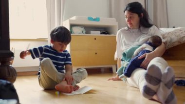 Anne oğlunu evde çizim yaparken izliyor, kucağında bebek, sıcak bir ev ortamında aile dinamiğini besliyor.
