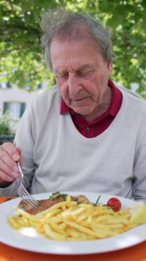 Açık bir kafede balık ve patates kızartması yiyen yaşlı bir adam, yeşil ve temiz havayla çevrili sakin bir ortamda yemek yemenin basit zevklerini vurguluyor. 