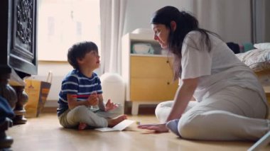 Anne ve oğlu yerde oturup sohbet ediyor ve bir çizim projesi üzerinde çalışıyorlar. Parlak ve rahat bir ev ortamında bir bağlantı ve yaratıcılık anı yaratıyorlar.