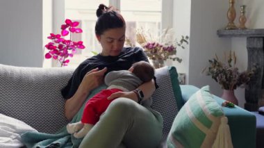 Anne bebeğini emzirirken parlak bir oturma odasındaki rahat bir koltukta, canlı bitkilerle çevrili ve rahat bir ortamda anne ile çocuk arasında huzurlu bir bağ olduğunu gösteriyor.