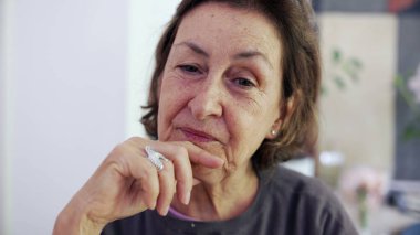 Üst düzey kadın, kamera önünde konuşulmayan birini dinliyor. Çenesinde el olan yaşlı bayan dikkatle dinliyor.