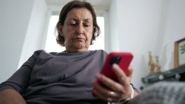 Oturma odasındaki kanepede otururken elinde cep telefonu cihazıyla içeriği okuyan yaşlı bir kadın. Ekrana bakan 70 'lerin ciddi ifadesi.