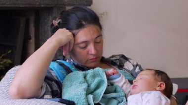 Uyuyan bebeğini kanepede tutan yorgun bir anne, başını elinde dinlendiriyor, erken anneliğin yorgunluğunu ve hassas anlarını gösteriyor.